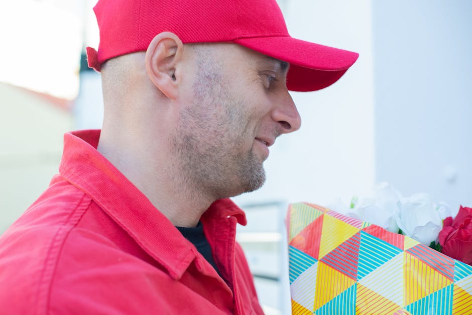 A man wearing a red baseball cap and red jacket is smiling gently while holding a colourful gift bag decorated with geometric patterns in shades of yellow, blue, red, and white, which contains white a