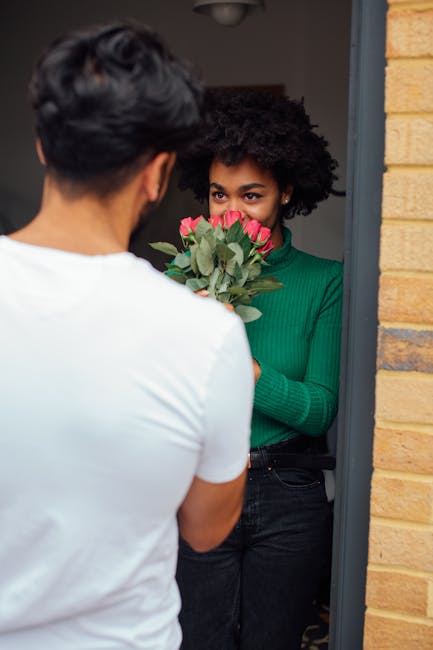 A woman with curly black hair wearing a green long-sleeve top is standing in a doorway, holding a bouquet of fresh pink roses with green foliage, ready for flower delivery. She is looking at a person